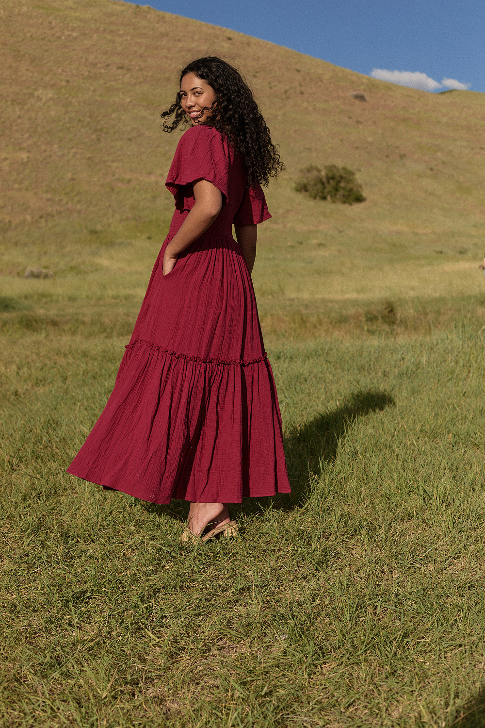 A woman with curly hair smiles over her shoulder on a grassy hill, wearing the Ivy City Co Lennon Maxi Dress in Wildberry—a flowing, berry-pink style that’s both nursing and maternity friendly. Green hills and a blue sky brighten the scene.