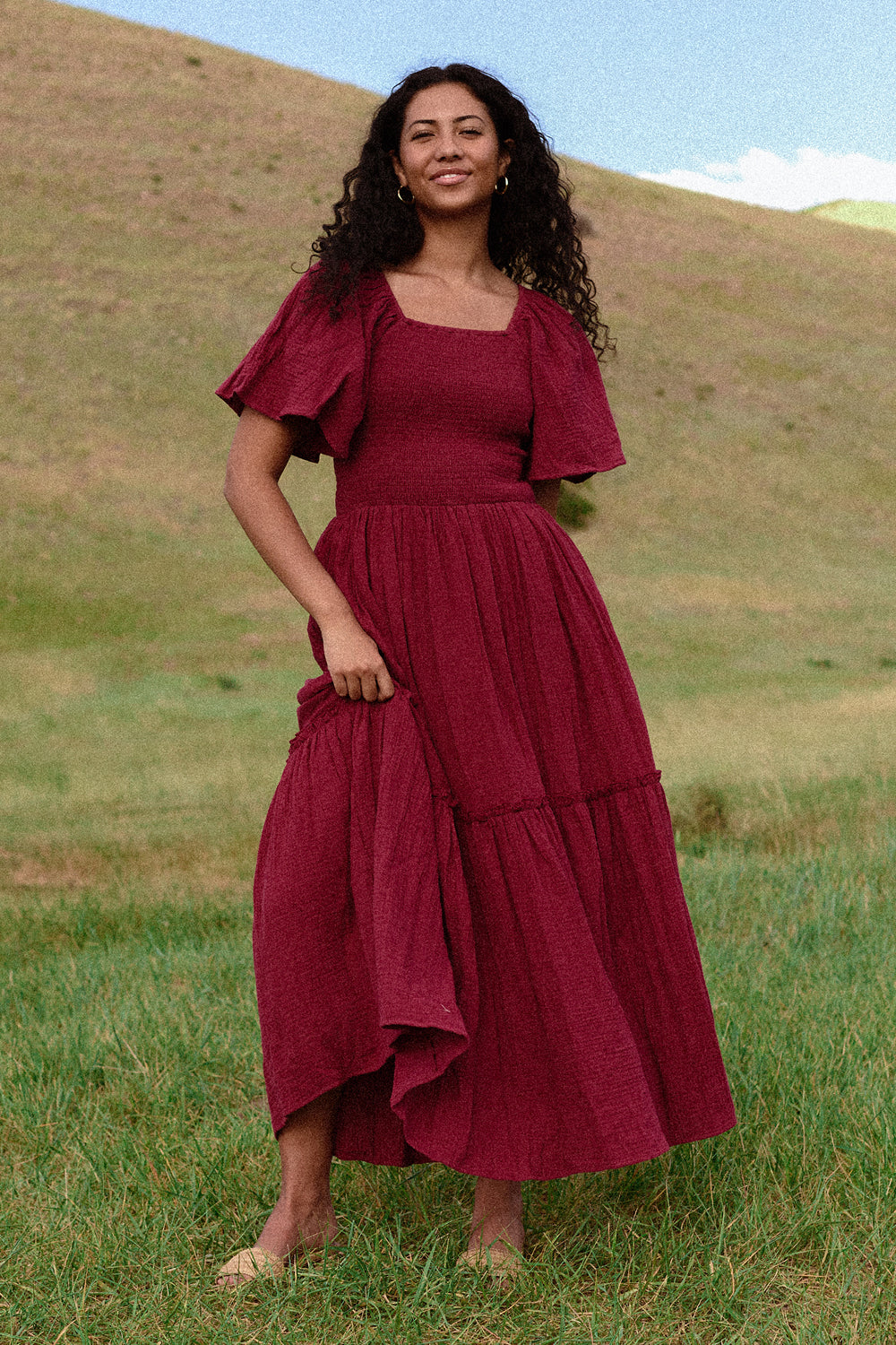 A woman with long curly hair stands barefoot on grass, smiling in the Ivy City Co Lennon Maxi Dress in Wildberry—a berry-pink, nursing and maternity-friendly maxi. Rolling hills and a partly cloudy sky create a serene backdrop.