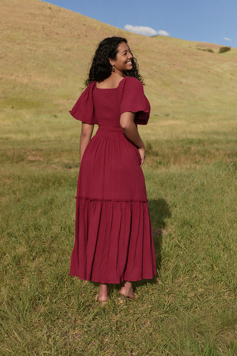 A woman with long curly hair stands barefoot in a grassy field, wearing Ivy City Co’s Lennon Maxi Dress in Wildberry—a nursing and maternity-friendly style. She faces away, gazing over her shoulder at the rolling hills and blue sky.