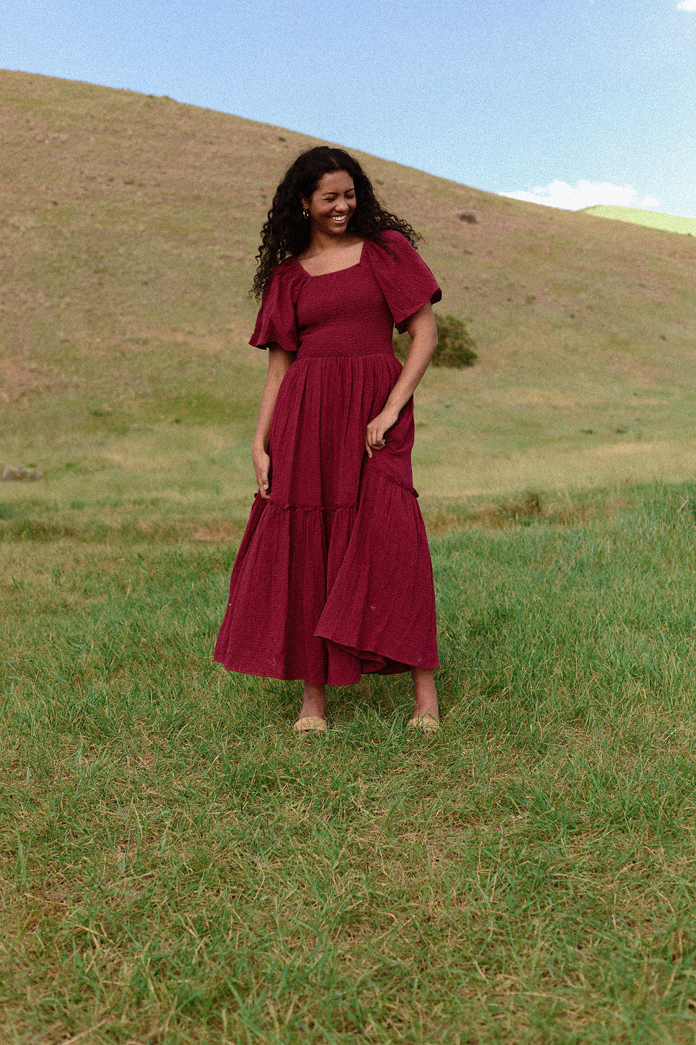 A woman wears the Ivy City Co Lennon Maxi Dress in Wildberry, smiling in a grassy field with rolling hills and blue sky behind her. The dress is both nursing and maternity friendly.