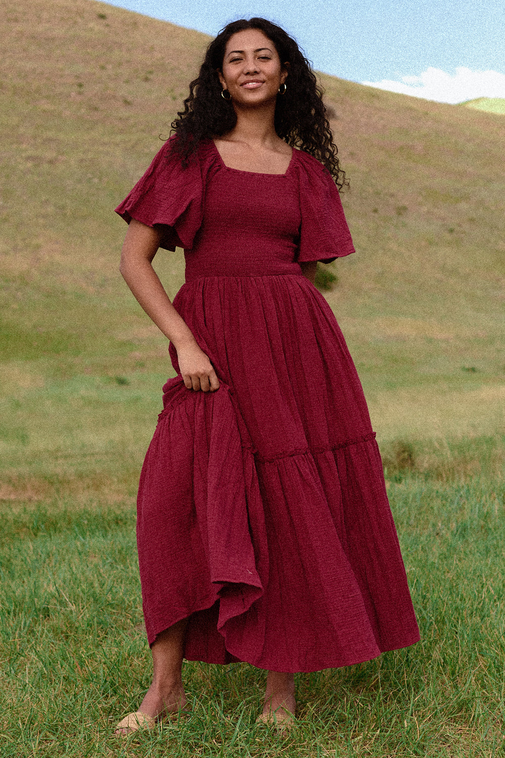 A woman with long curly hair stands on grass before a hillside, wearing the Ivy City Co Lennon Maxi Dress in Wildberry—flowing with short flutter sleeves, nursing and maternity friendly. The sky above is blue with wispy clouds.