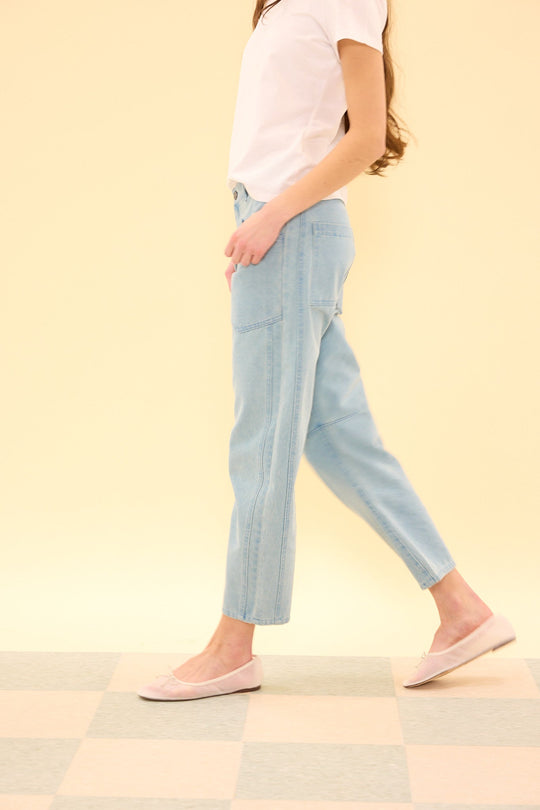 An individual wearing a white t-shirt, Ivy City Co’s Amanda Pants in Light Wash Denim, and pink flats walks on a checkered floor with a pale yellow background. Their face is not visible.