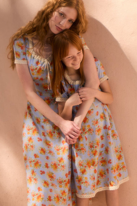 Two people with long red hair stand close together, both smiling warmly in soft sunlight. They wear matching Ivy City Co Mini Libby Dresses in Orange Floral against a peach-colored wall. The taller one hugs the smaller lovingly.