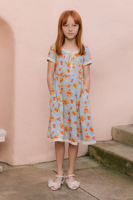 A young girl with long red hair stands on a concrete surface in front of a pale pink wall, wearing the Ivy City Co Mini Libby Dress in Orange Floral and white bow sandals. Her hands are in her pockets as she looks at the camera.