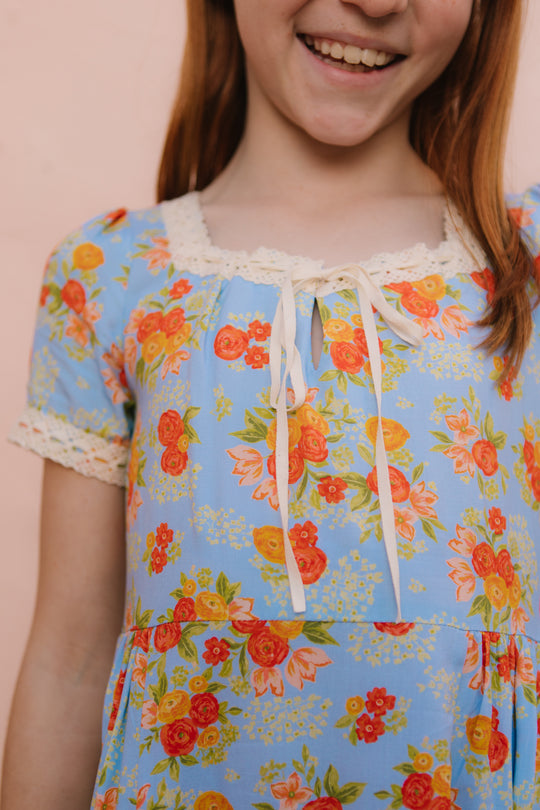 A girl smiles, wearing the Ivy City Co Mini Libby Dress in Orange Floral. The blue midi-length dress features a floral print and white lace trim on the collar and sleeves. The photo is cropped from the shoulders down.