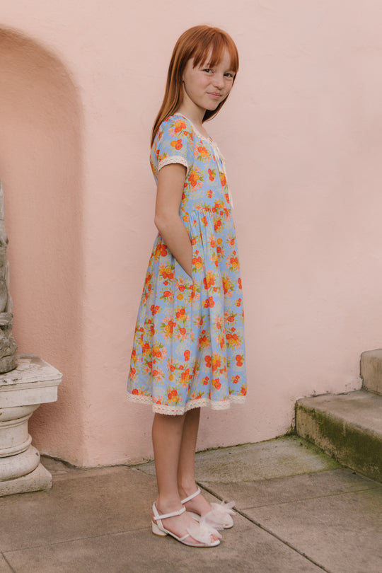 A young girl with straight red hair stands sideways outdoors by a pale pink wall and stone steps, wearing Ivy City Cos Mini Libby Dress in Orange Floral with white sandals.