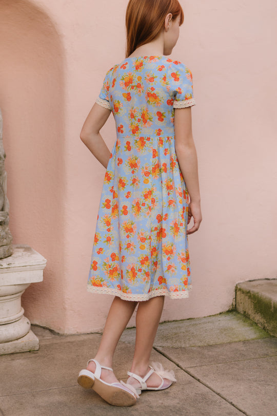 A young girl in the Ivy City Co Mini Libby Dress in Orange Floral stands on a stone path facing away, next to a pale pink wall and stone bench, wearing white sandals. The dress features a light blue base with orange floral print and white trim.