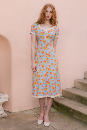A woman with wavy red hair wears the Ivy City Co Libby Dress in Orange Floral—an ankle-length, light blue dress with orange florals and short sleeves—standing by outdoor stone steps against a pale peach wall, paired with white shoes.