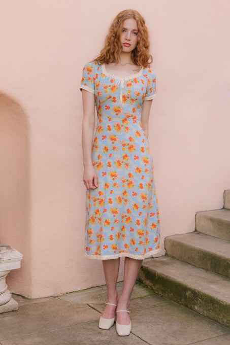 A woman with wavy red hair wears the Ivy City Co Libby Dress in Orange Floral—an ankle-length, light blue dress with orange florals and short sleeves—standing by outdoor stone steps against a pale peach wall, paired with white shoes.