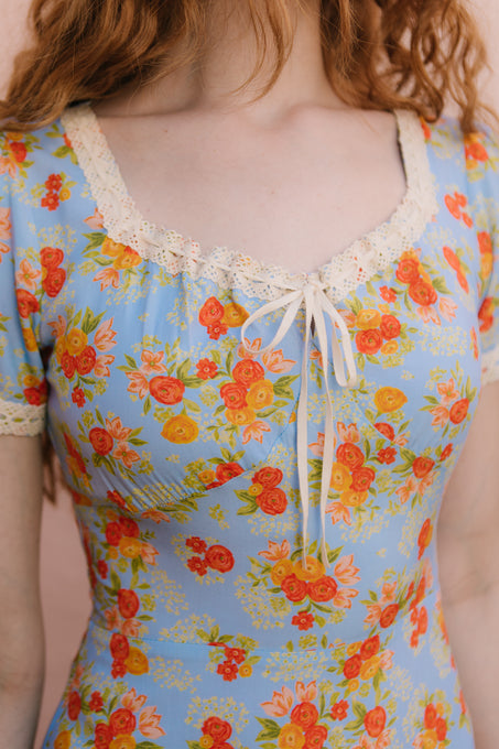 A woman with long red hair wears the Libby Dress in Orange Floral by Ivy City Co, featuring red and yellow flowers, cream crochet lace at the neckline and sleeves, and a small bow accent at the neckline.