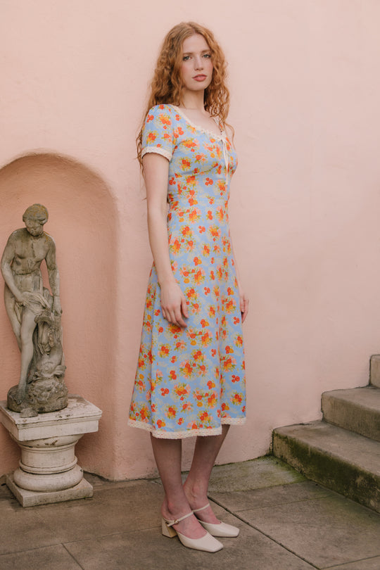 A woman with long red hair in an Ivy City Co Libby Dress in Orange Floral and cream heels stands on a stone patio near steps and a statue, against a pale pink wall.