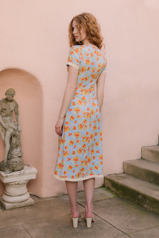 A woman with wavy red hair stands on a stone patio beside a classical statue, wearing the Ivy City Co Libby Dress in Orange Floral and high-heeled shoes.