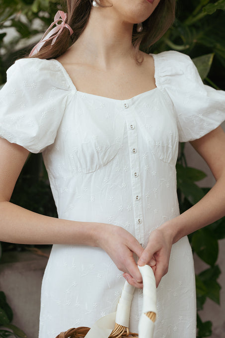 A woman wears the Liberty Dress in White Eyelet by Ivy City Co, featuring puffed sleeves and front buttons, and holds a white-handled basket. She has a pink ribbon in her hair and stands before lush green plants.