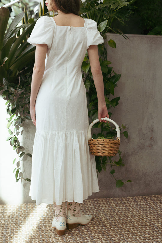 A woman faces away, holding a wicker basket, in the Ivy City Co Liberty Dress in White Eyelet with puff sleeves and a fit and flare silhouette. She pairs it with white floral-embroidered boots, amid green plants by a textured wall.