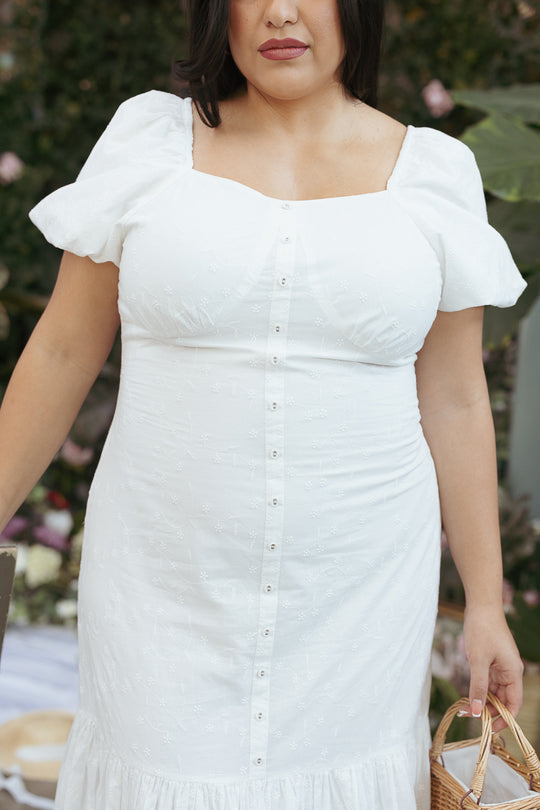 A woman stands outdoors amid greenery, holding a small woven basket and wearing the Ivy City Co Liberty Dress in White Eyelet. The cropped photo (mouth to knees) highlights the fit and flare silhouette with short sleeves.
