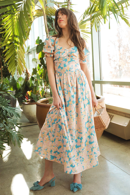 A woman wears the Ivy City Co Mila Dress in Floral, featuring puff sleeves, indoors among large green plants. She holds a woven handbag, gazes upward as sunlight streams through tall windows, and sports blue bow shoes.