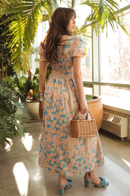 Indoors among green plants, a woman wears the Ivy City Co Mila Dress in Floral with puff sleeves and blue shoes. Holding a wicker handbag, she looks over her shoulder as sunlight streams through large windows behind her.