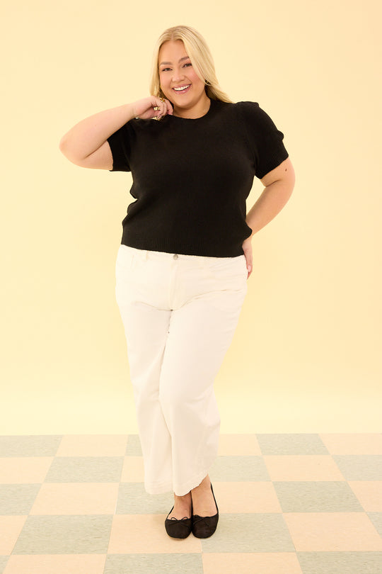 Woman wearing a black top and white pants on a checkered floor with a beige wall background