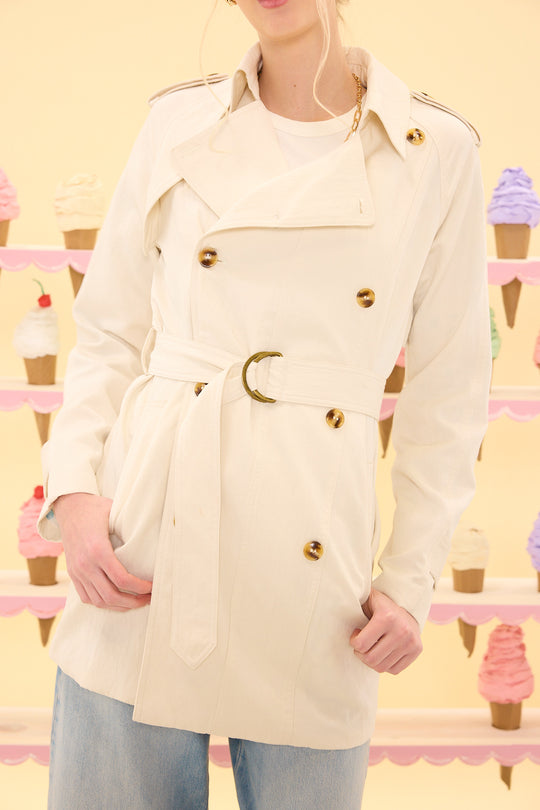 Person wearing a cream-colored trench coat with gold buttons against a pastel ice cream cone background.