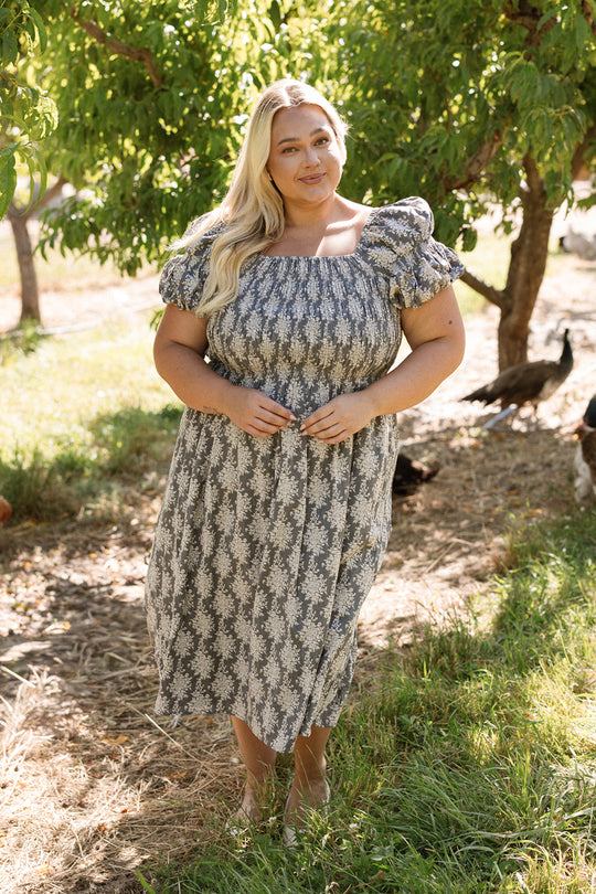 A woman with long blonde hair stands outdoors, gently smiling in the Lisa Dress in Jolie Fleur by Ivy City Co. The smocked bodice offers a flattering and comfortable fit, making this style both elegant and maternity friendly. She is surrounded by green trees and sunlight, with grass and a few birds in the background.