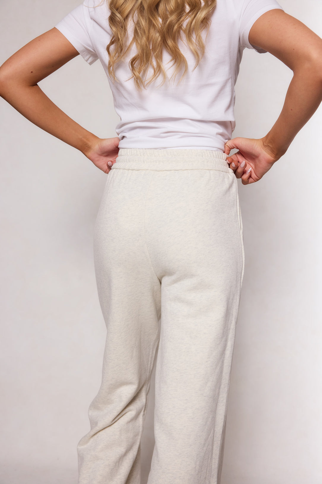 A woman stands with hands on hips, shown from the back, wearing Ivy City Co’s Lisbeth Sweatpant in Oatmeal and a white t-shirt, set against a plain light background.