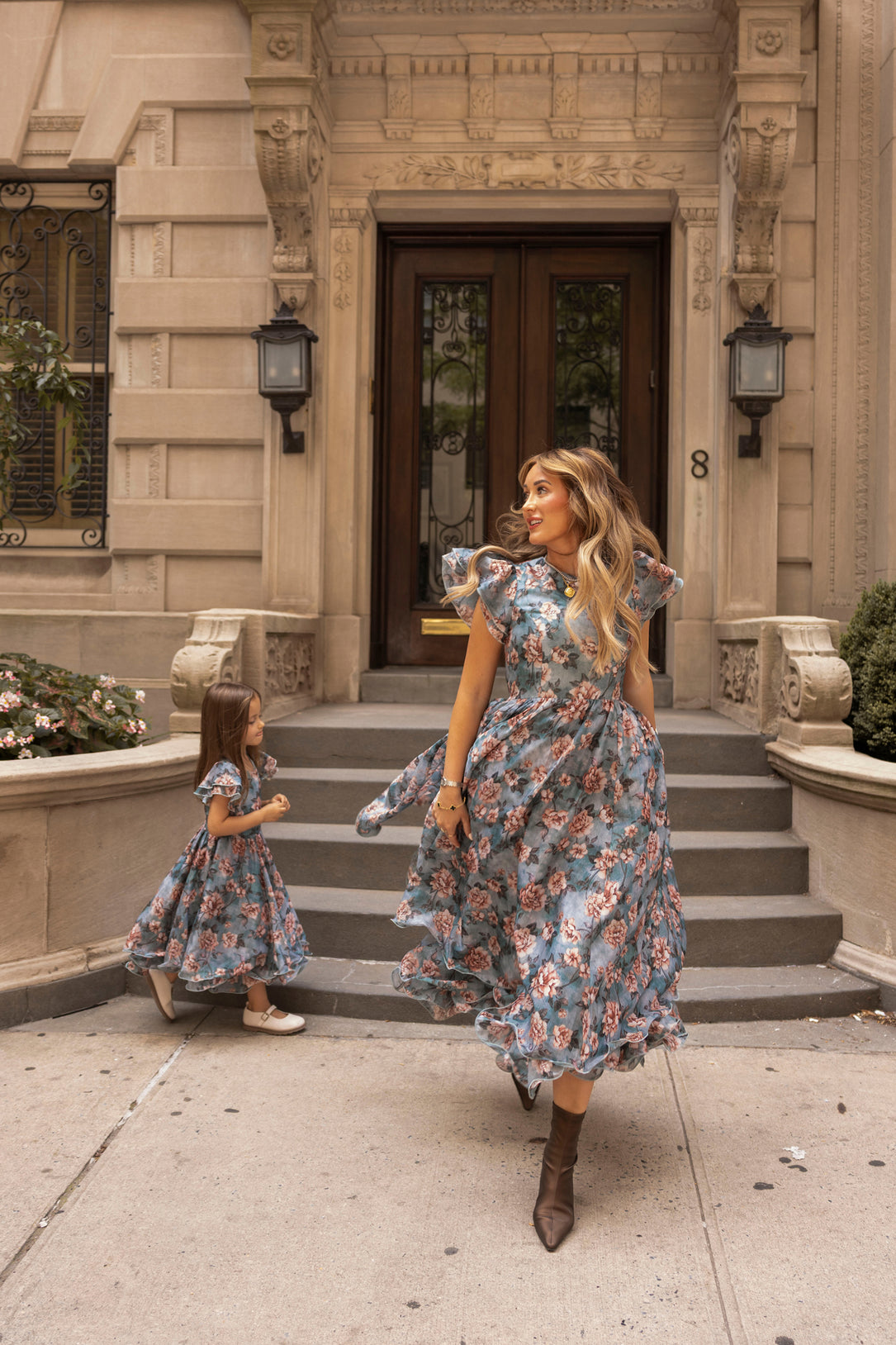 A woman and a young girl, both wearing the Ivy City Co Lisette Dress in Rose Dusk, walk in front of a grand building with stone steps and arched doors. The woman looks back at the girl, who follows behind.