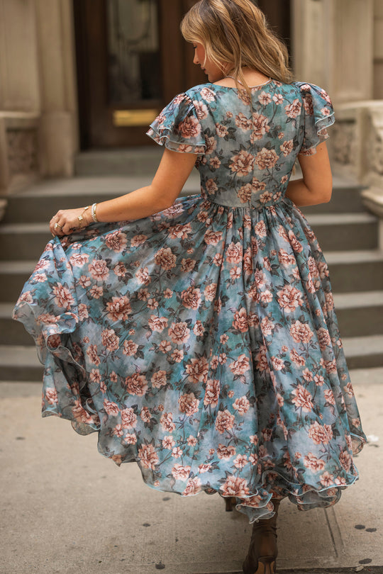 A woman twirls on a city sidewalk in the Rose Dusk Lisette Dress by Ivy City Co, holding out the skirt to show off the tea-length dress’s flowy fabric and flutter sleeves. She faces away, with stone steps and a building in the background.