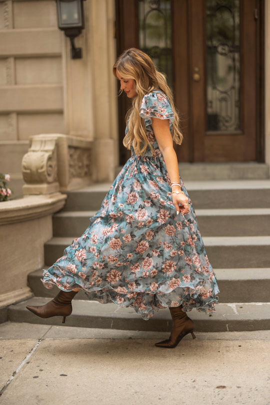 A woman in a flowing, floral Lisette Dress in Rose Dusk by Ivy City Co and brown heeled boots walks gracefully on a city sidewalk, stepping down a concrete curb near a stone building with ornate details.