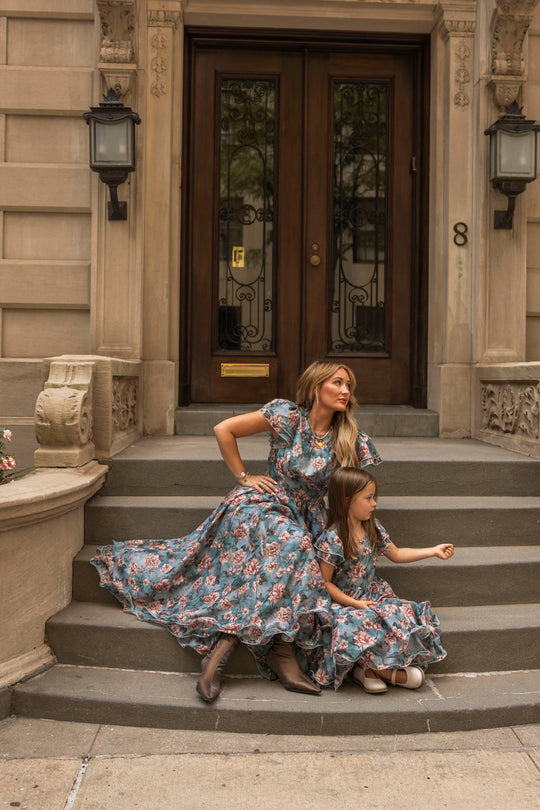 A woman and a young girl, both dressed in matching Ivy City Co Lisette Dresses in Rose Dusk, featuring floral patterns and tea-length hems, sit on stone steps before the grand wooden door of a classic building. The woman gazes away while the girl sits quietly beside her.