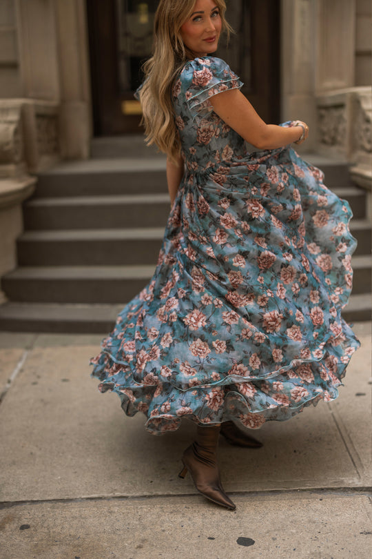 A woman twirls in the flowy Rose Dusk Lisette Dress by Ivy City Co and brown boots on a city sidewalk, with stone steps and a building entrance in the background.