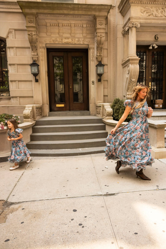 A woman and a young girl, both wearing the Ivy City Co Lisette Dress in Rose Dusk, stroll and dash in front of a stone building with steps and ornate details.