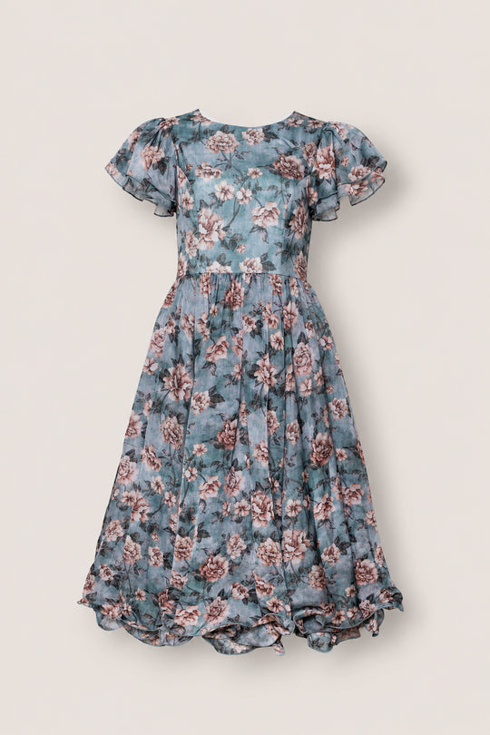 The Lisette Dress in Rose Dusk by Ivy City Co is a blue tea-length dress adorned with a charming pink and white floral pattern. Designed with short flutter sleeves and a flared, ruffled hem, this elegant dress is an ideal choice for any occasion.
