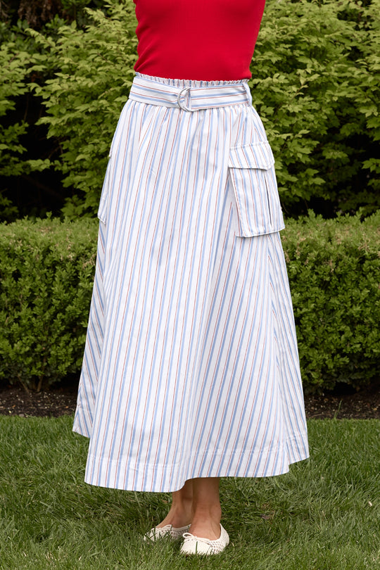 Someone wearing a red top and the Ivy City Co Lizzy Skirt in Heritage Stripe with large side pockets stands on grass in front of green bushes, their face not visible.