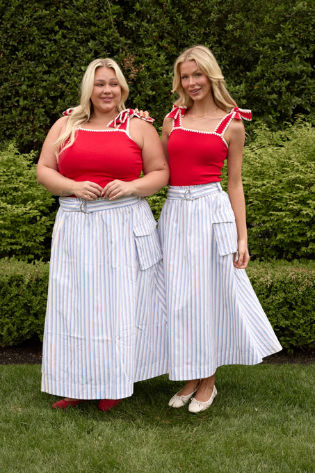 Two women smile at the camera in matching red sleeveless tops and Ivy City Co’s Lizzy Skirt in Heritage Stripe, a perfect summer style with large pockets. They stand on grass before bushes, looking cheerful—one has blonde hair.