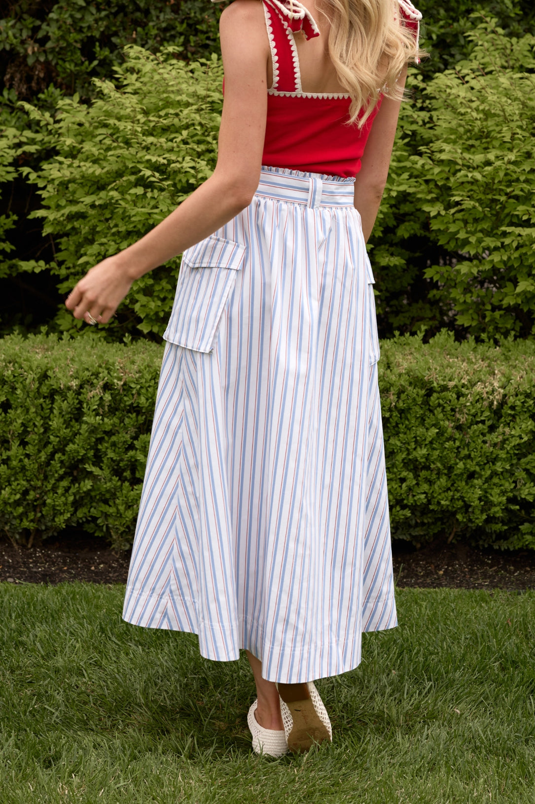 A woman, back to the camera, stands on grass wearing a red sleeveless top and Ivy City Co’s Lizzy Skirt in Heritage Stripe—a high-waisted summer skirt with blue and orange stripes and large side pockets. Green bushes are in the background.