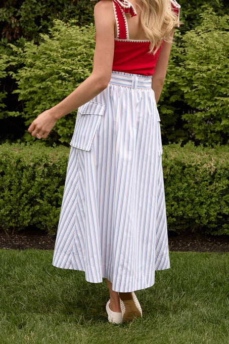 A woman, back to the camera, stands on grass wearing a red sleeveless top and Ivy City Co’s Lizzy Skirt in Heritage Stripe—a high-waisted summer skirt with blue and orange stripes and large side pockets. Green bushes are in the background.