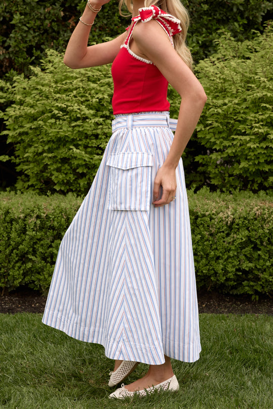 A woman stands outside in a red sleeveless top and the Ivy City Co Lizzy Skirt in Heritage Stripe—a blue-and-white, high-waisted summer skirt with a large pocket—paired with beige flats, green bushes behind her.