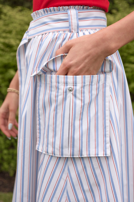 Wearing the Lizzy Skirt in Heritage Stripe by Ivy City Co—a blue and white midi with a large front pocket—a person stands outdoors among lush greenery, hand tucked in the pocket, paired with a red top and bracelet.