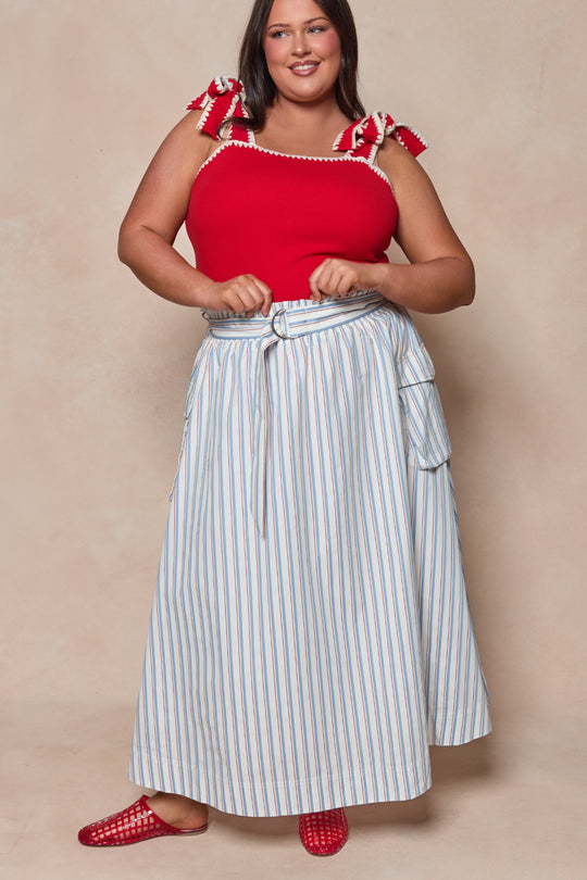 A woman smiles in red shoes and a red sleeveless top with bows, styled with the Ivy City Co Lizzy Skirt in Heritage Stripe—a belted blue-and-white summer midi—posing against a beige background.