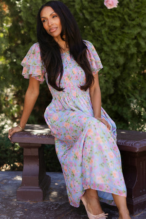 A woman with long dark hair sits on a brown bench outdoors, wearing the Ivy City Co Lottie Dress in Vibrant English Garden—a floral, maternity-friendly dress with ruffled sleeves—surrounded by greenery and sunlight.