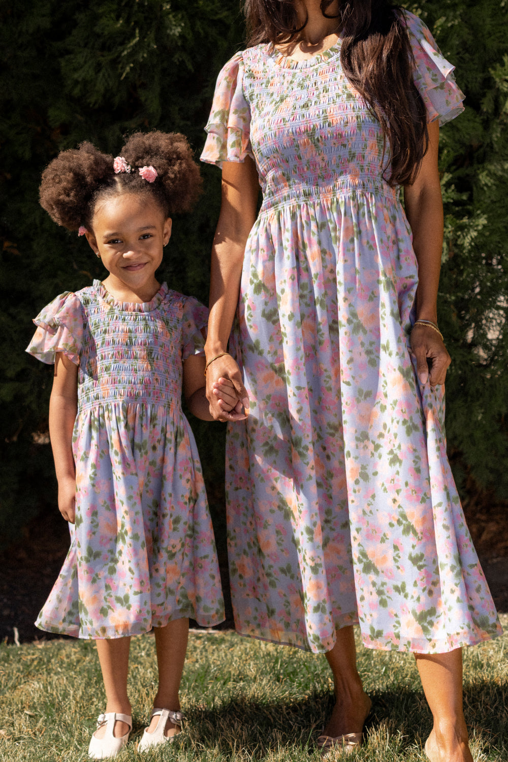 A smiling adult and child hold hands outdoors in matching Ivy City Co Lottie Dresses in Vibrant English Garden, featuring soft florals, ruffled sleeves, smocked tops, and a maternity-friendly fit. The child’s afro puffs are styled with pink bows.