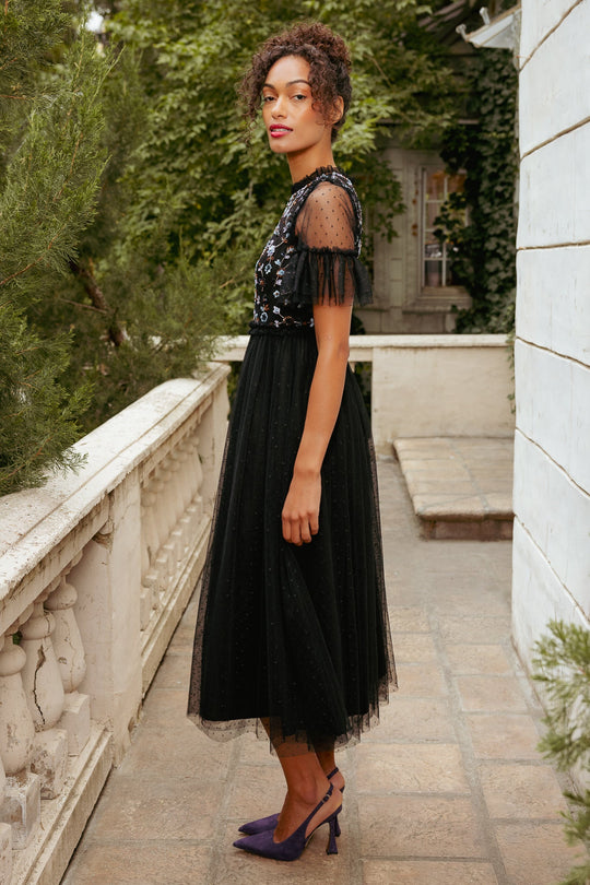 A woman stands sideways on a stone balcony, wearing the Ivy City Co Louisa Dress in Black—a tulle dress adorned with delicate floral embroidery—and purple heels, surrounded by greenery and white walls.