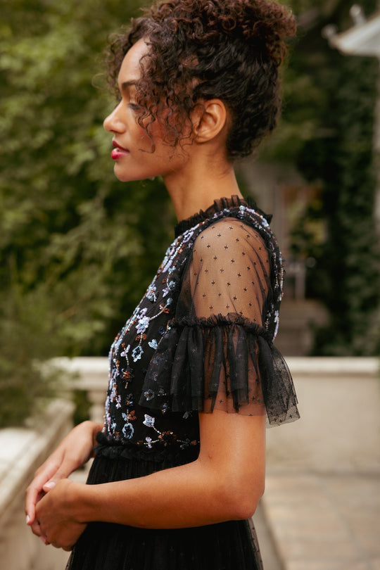 A woman with curly hair in a bun wears the Louisa Dress in Black by Ivy City Co—a tulle dress adorned with sheer ruffled sleeves and delicate floral embroidery—while standing outdoors amidst lush greenery and stone steps.