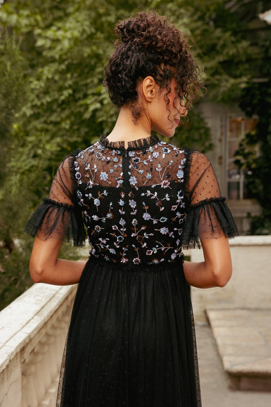 A woman with curly hair stands on a balcony, surrounded by greenery, wearing the Louisa Dress in Black by Ivy City Co—a sheer tulle gown adorned with blue floral embroidery. She faces away from the camera.