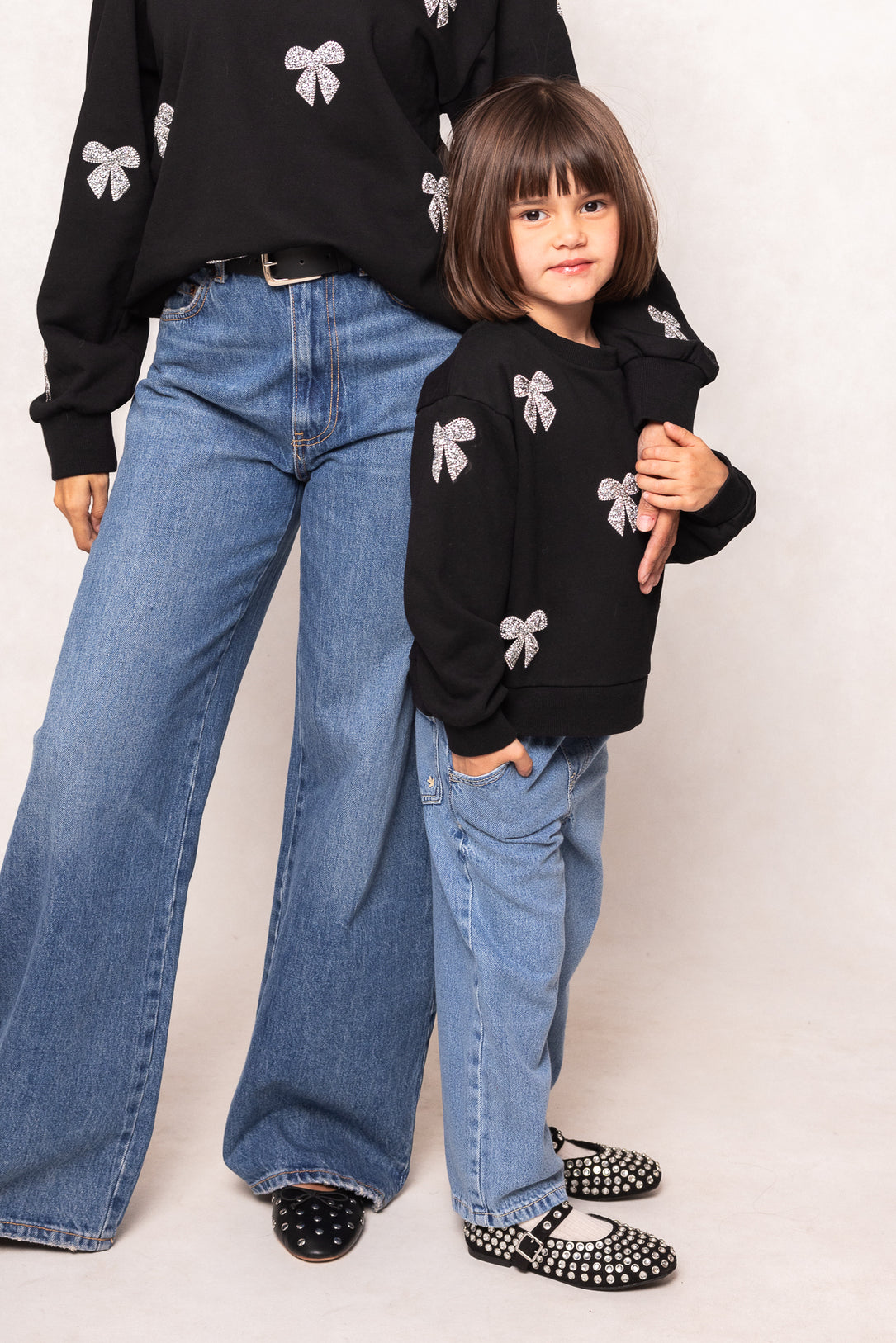 Two people wearing matching black sweaters with white bow designs and blue jeans on a plain background.