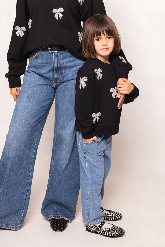 Two people wearing matching black sweaters with white bow designs and blue jeans on a plain background.
