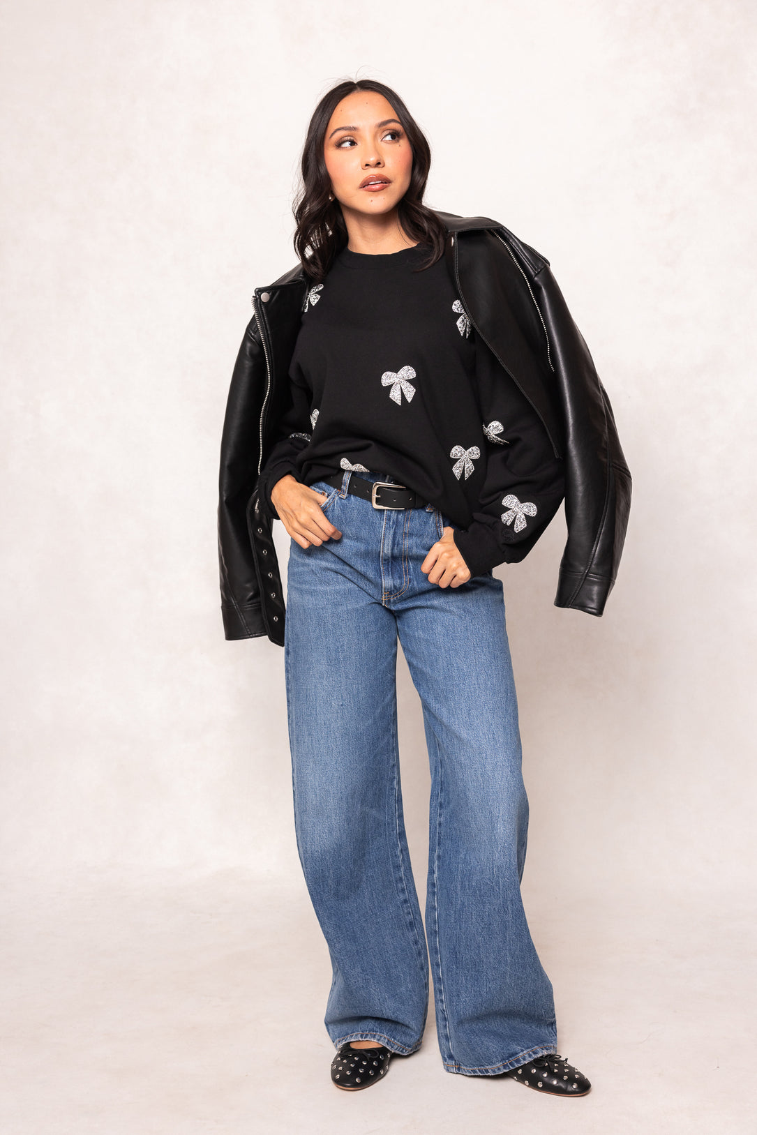 Woman wearing a black leather jacket, black floral sweater, and blue jeans on a white background