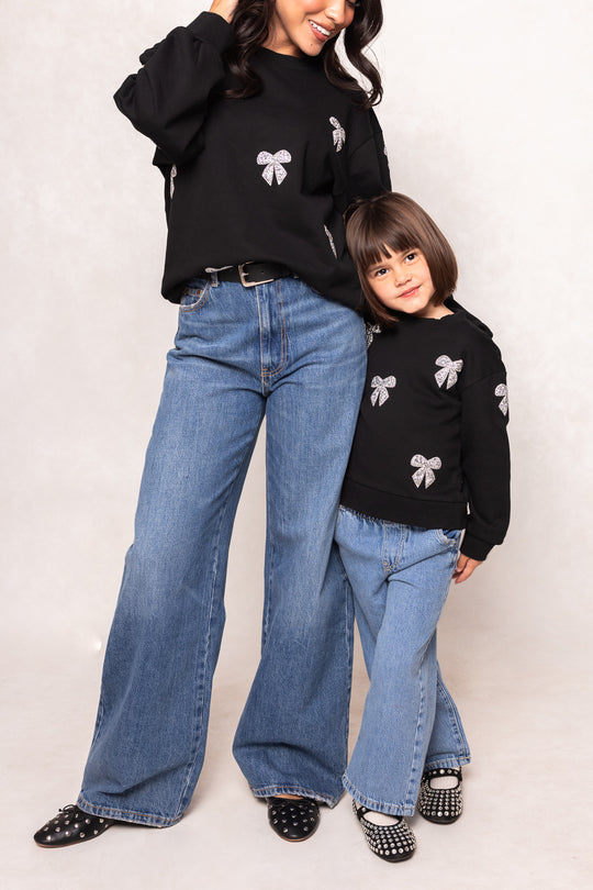 Woman and child wearing matching black sweaters with white patterns and blue jeans on a plain background