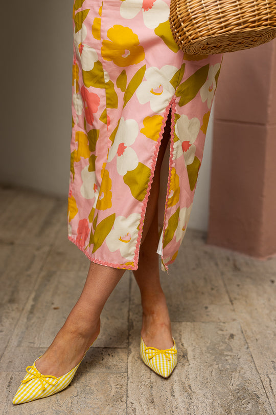 A person in the Ivy City Co Lula Dress Made With Liberty Fabric, featuring a pink, white, and yellow floral print with a front slit, stands on a stone floor. They wear yellow checkered pointed-toe flats and carry a wicker bag. The image is cropped at the knee.