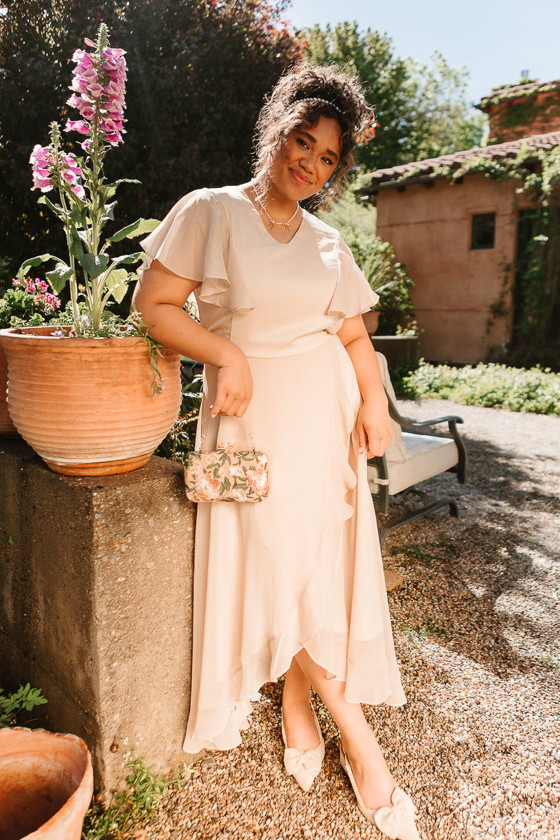 A woman stands outdoors near blooming flowers on a sunny day, smiling at the camera as she holds a small floral bag and wears the Callie Dress in Champagne Chiffon by Ivy City Co.